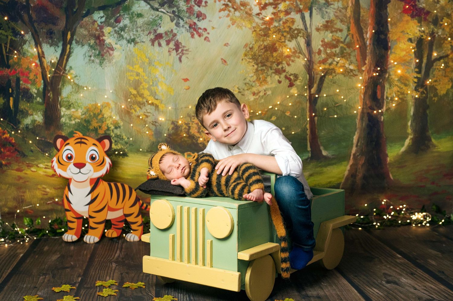 Boy and sleeping baby in tiger costume pose in toy jeep with stuffed tiger on woodland backdrop.