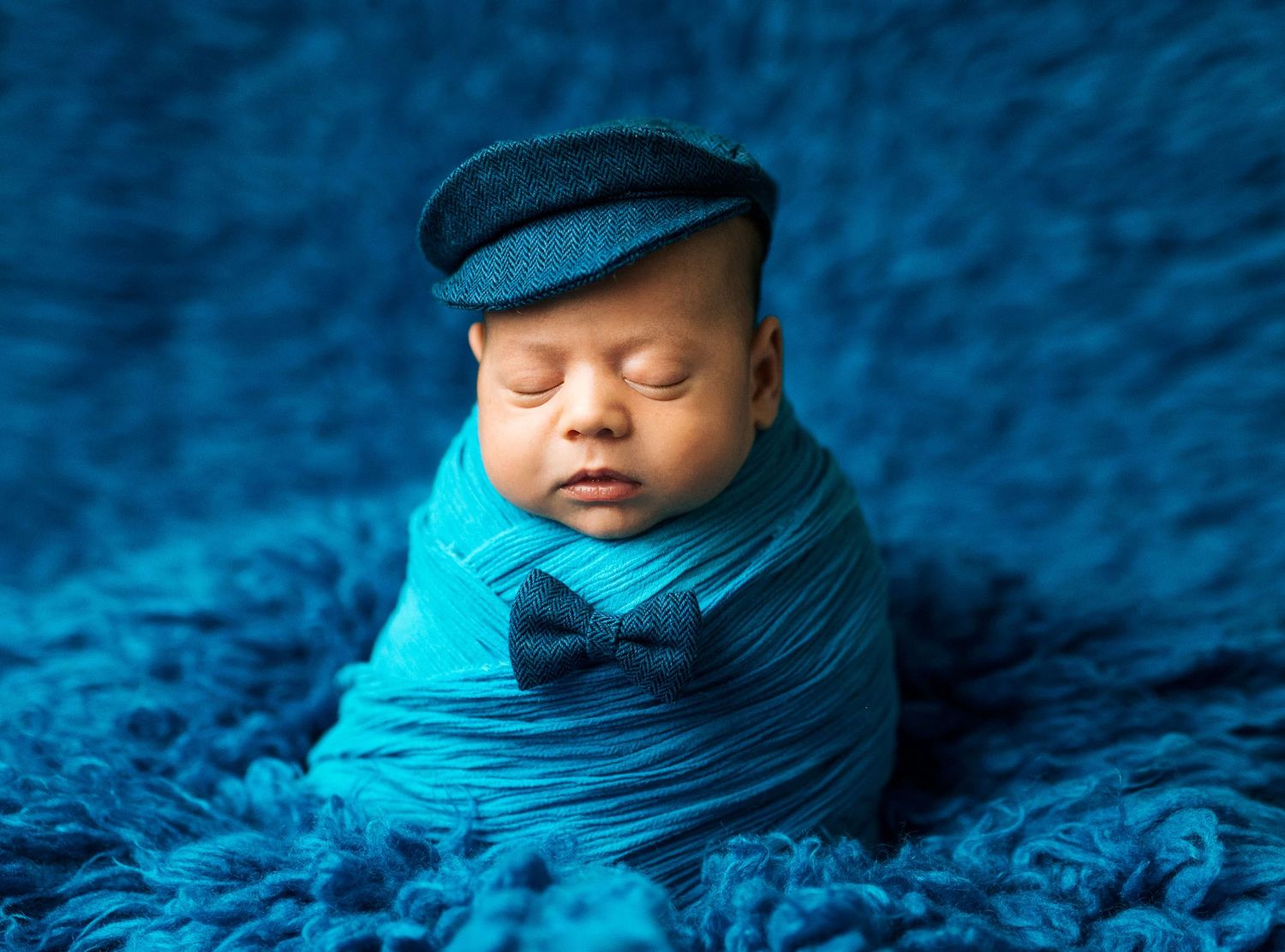 Newborn baby wrapped in teal fabric wearing a tiny flat cap and bow tie on a blue fluffy background.