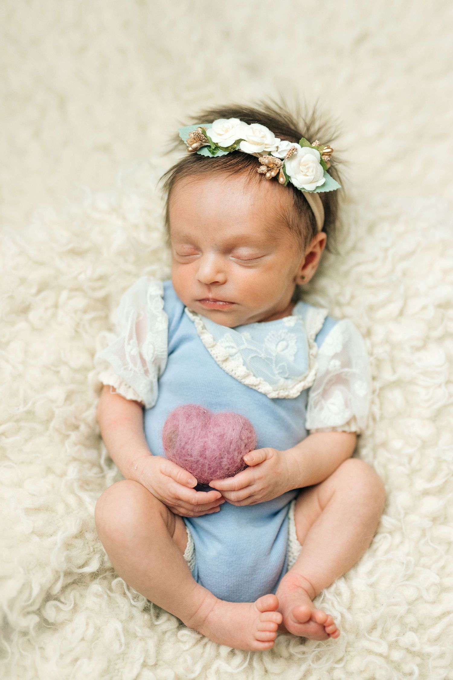 Sleeping newborn baby girl in blue romper holding pink heart, wearing floral crown, on cream fluffy blanket.