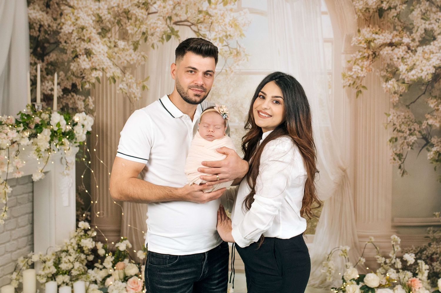 A couple poses with their newborn baby amid elegant floral decor in a professional studio portrait.
