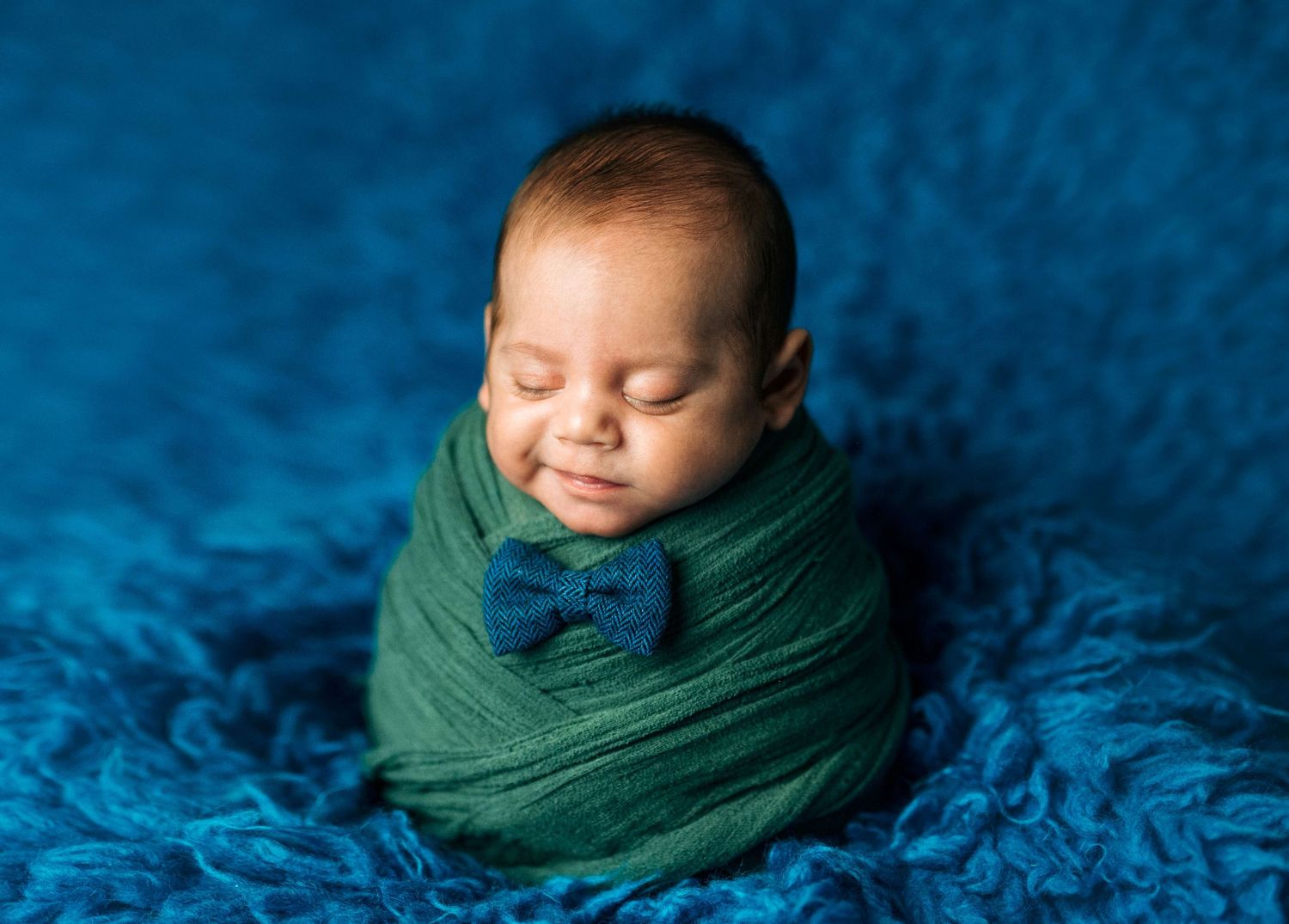 Smiling newborn baby swaddled in green wrap with blue bow tie on blue fluffy blanket.