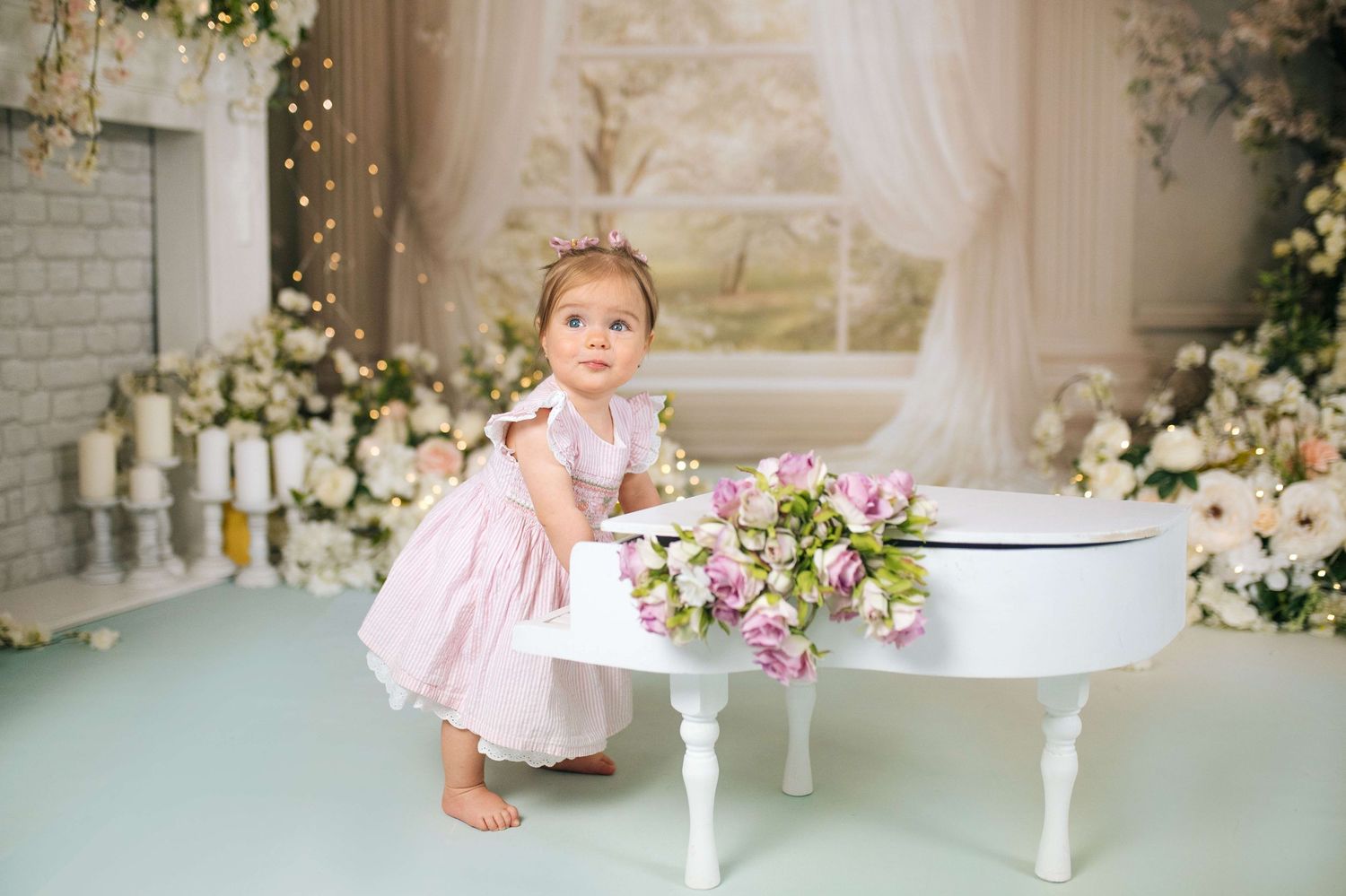 Toddler girl in pink dress poses beside a white mini grand piano with flowers in a decorated studio.