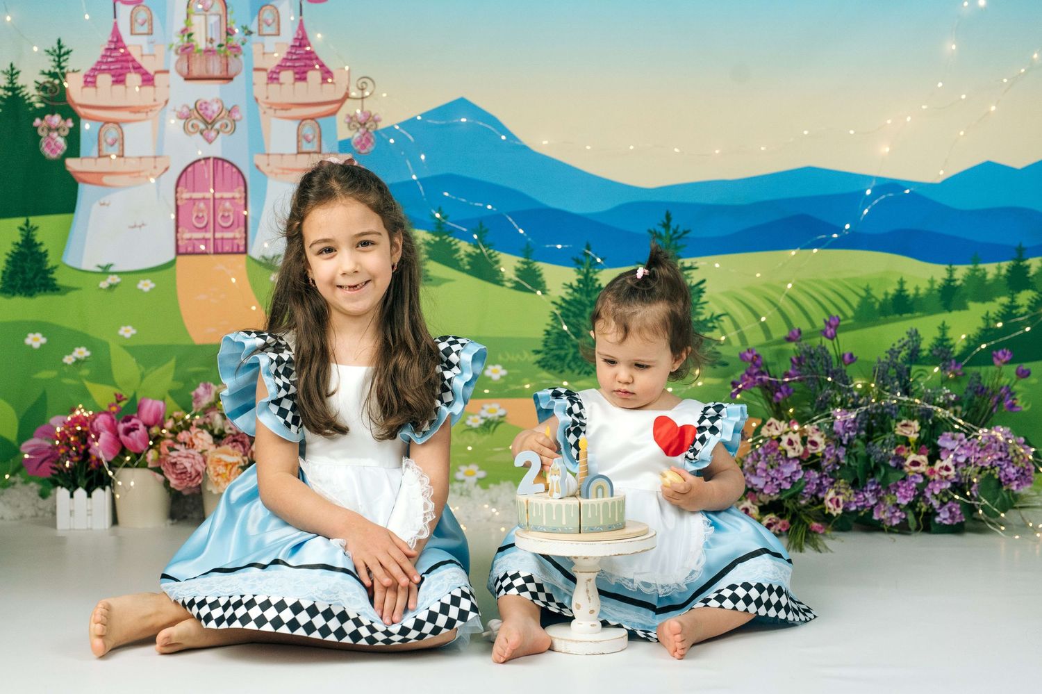 Two young girls in Alice in Wonderland dresses pose with a birthday cake against a fairy tale castle backdrop.