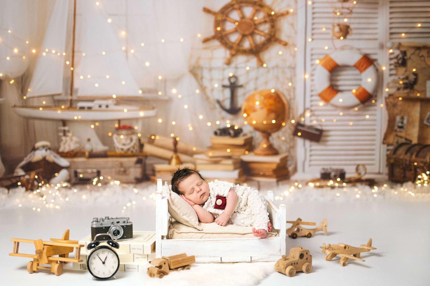 Newborn baby sleeping in tiny white bed surrounded by wooden toy planes, nautical decor, and warm fairy lights.