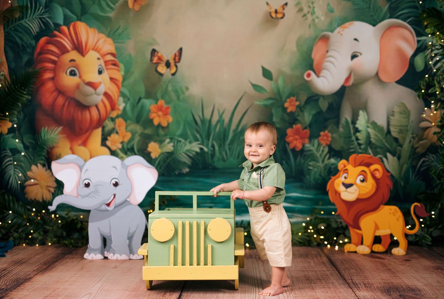 Smiling baby boy stands beside a toy jeep in a jungle safari themed photo backdrop with cartoon animals.