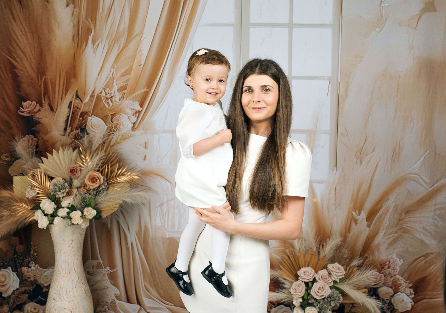 Woman in white dress holds toddler in white outfit, posing before boho floral backdrop with pampas grass decor.