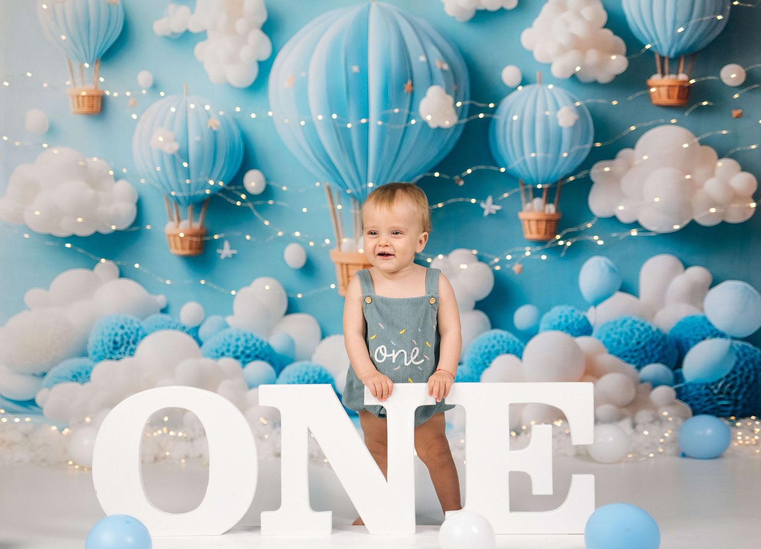 Baby's first birthday cake smash with blue hot air balloon backdrop, white ONE letters, and festive balloon decorations.