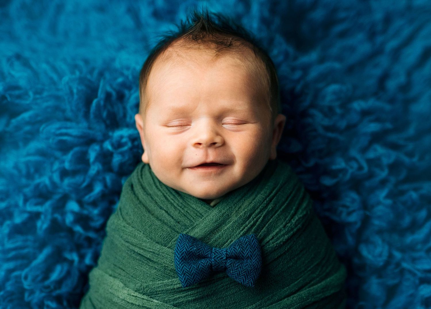 Smiling newborn baby swaddled in green wrap with blue bow tie on teal fluffy background.