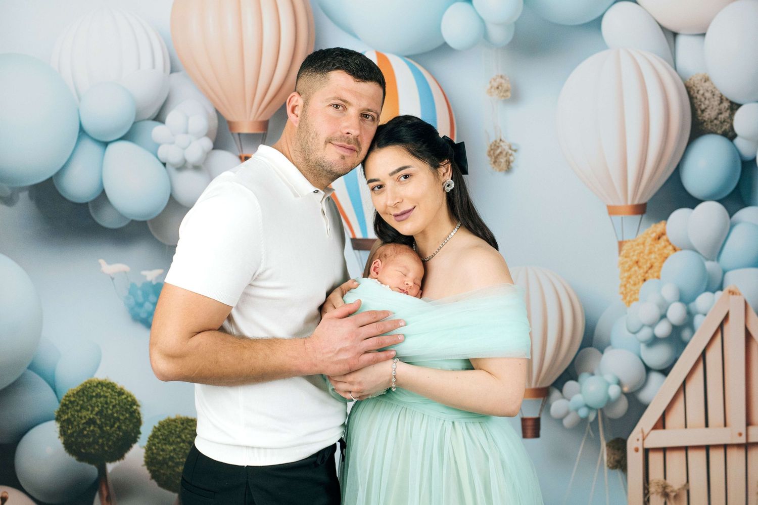 A couple poses with their newborn baby in front of a blue balloon and hot air balloon themed backdrop.