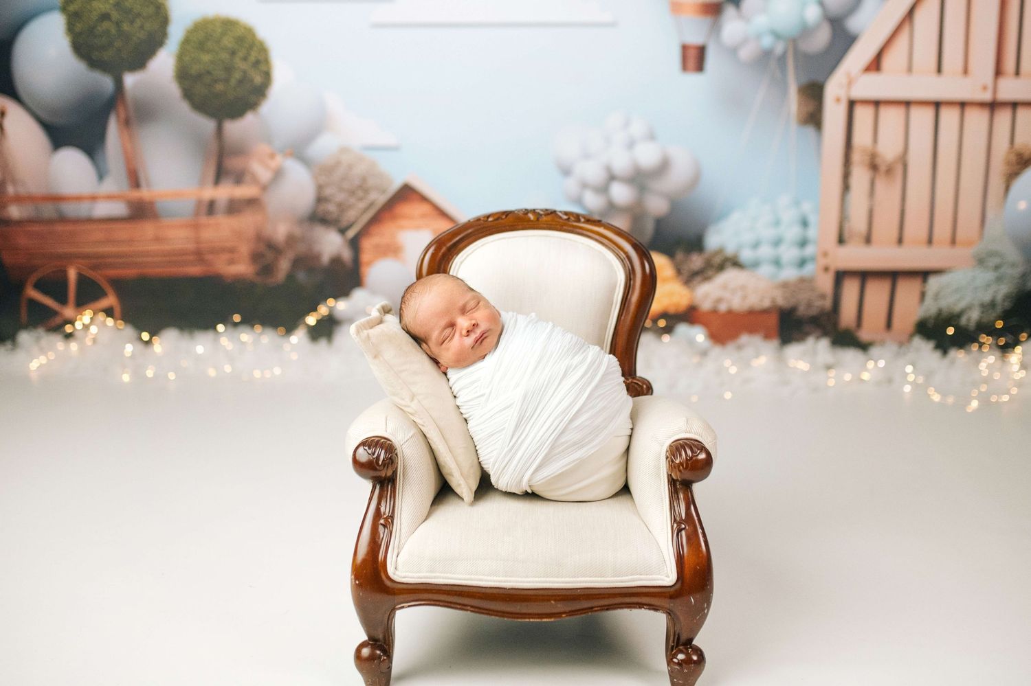 Newborn baby swaddled in white wrap sleeping peacefully in a miniature cream armchair with fairy lights and balloon backdrop.