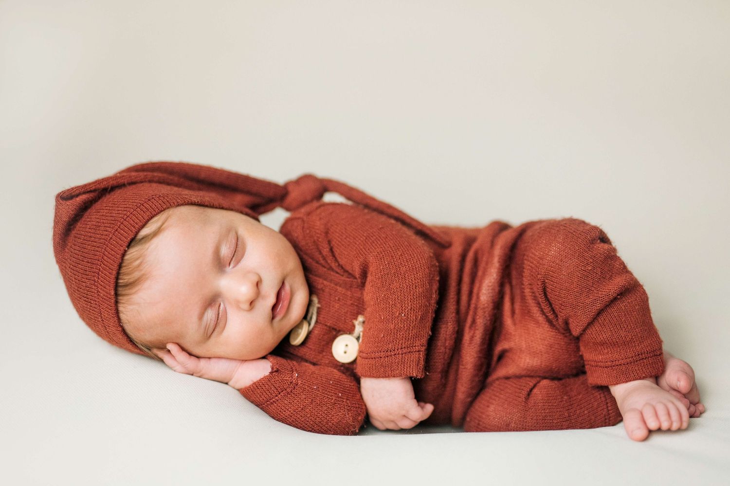 Sleeping newborn baby in rust-colored knit outfit and matching bonnet hat posed on white background.