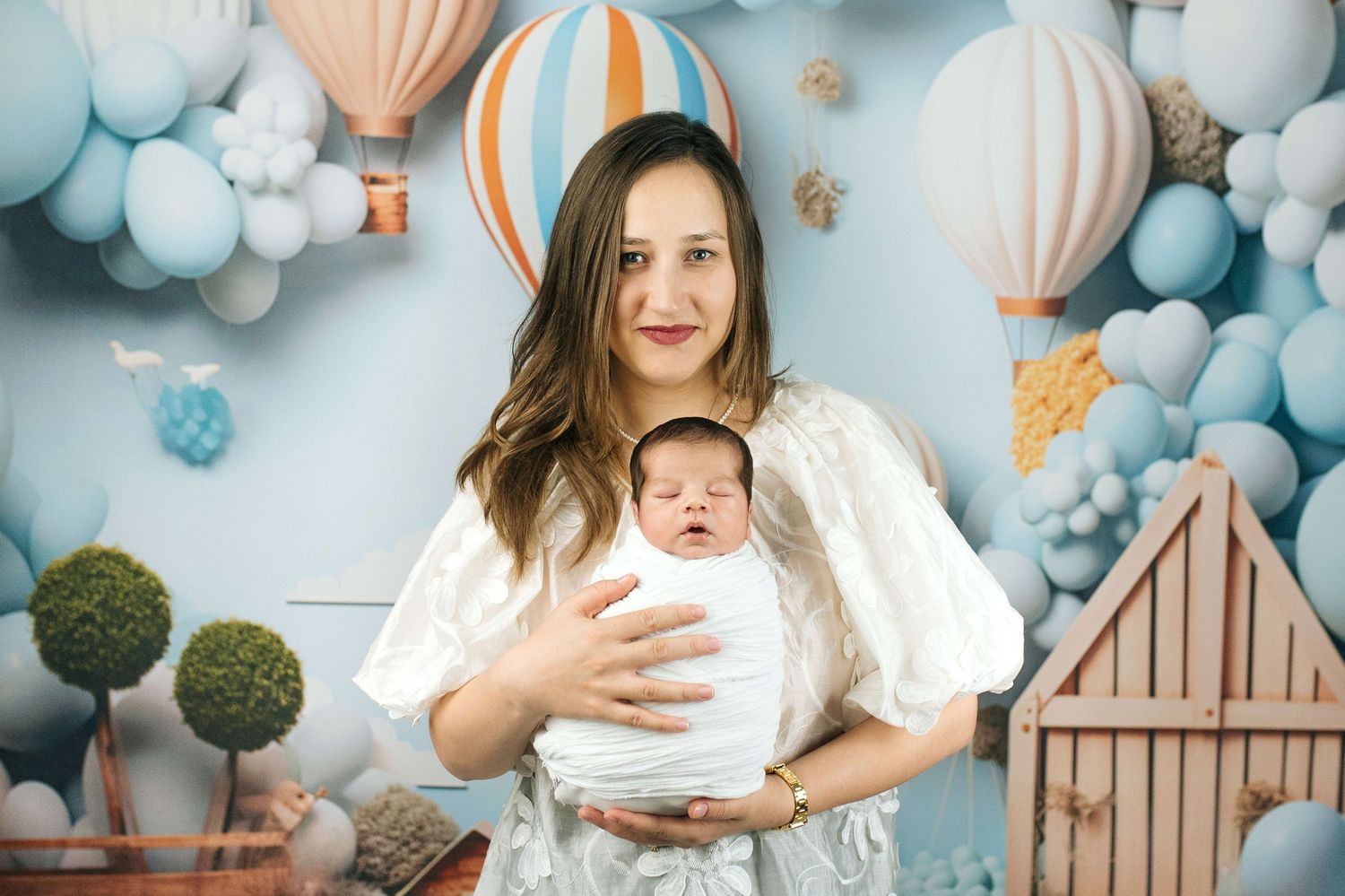Mother holding newborn baby in white wrap against blue balloon and hot air balloon themed backdrop.