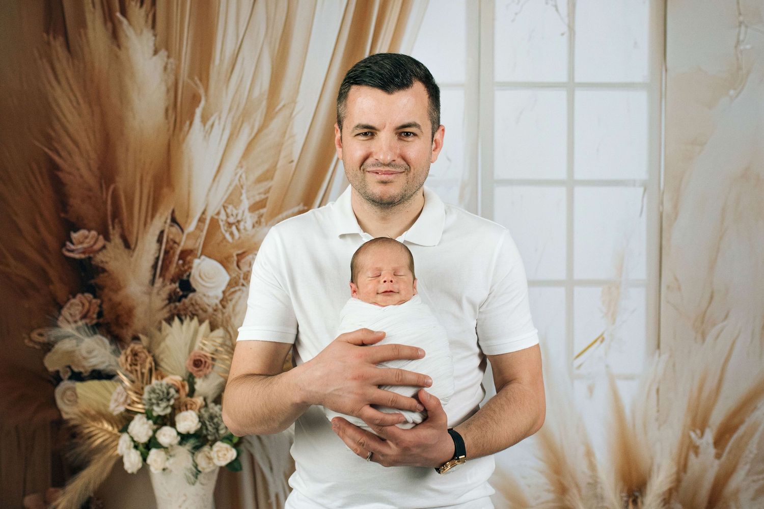 Man in white polo shirt proudly holds sleeping newborn baby in studio with boho floral decor.