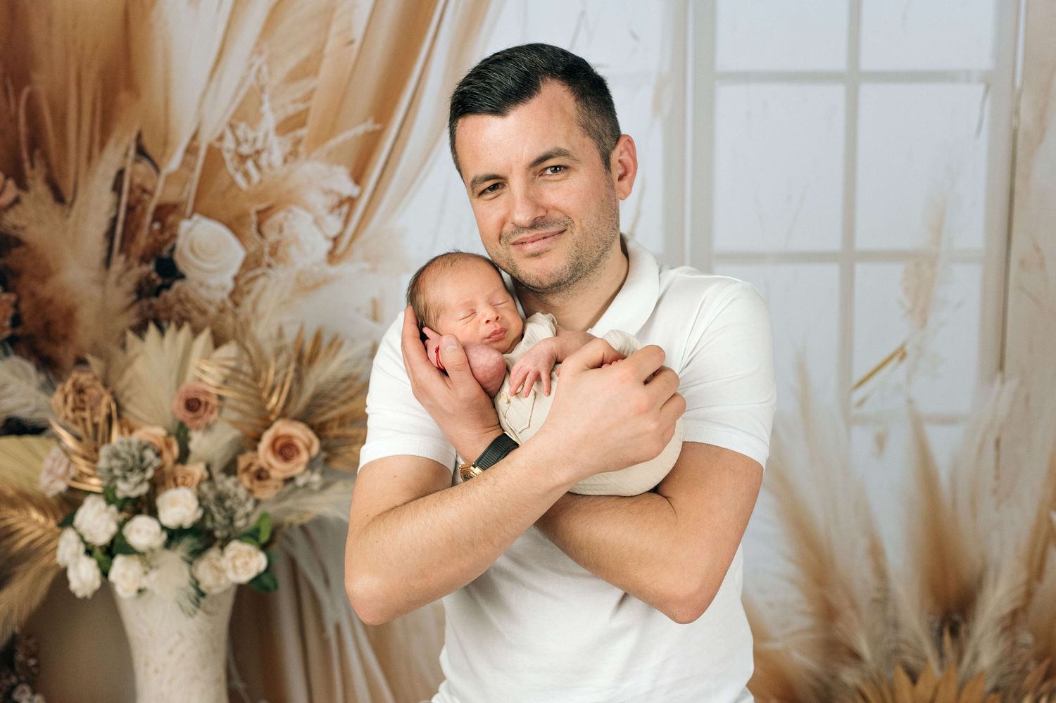 A smiling father in a white polo shirt gently holds his sleeping newborn baby against his chest in a studio setting.