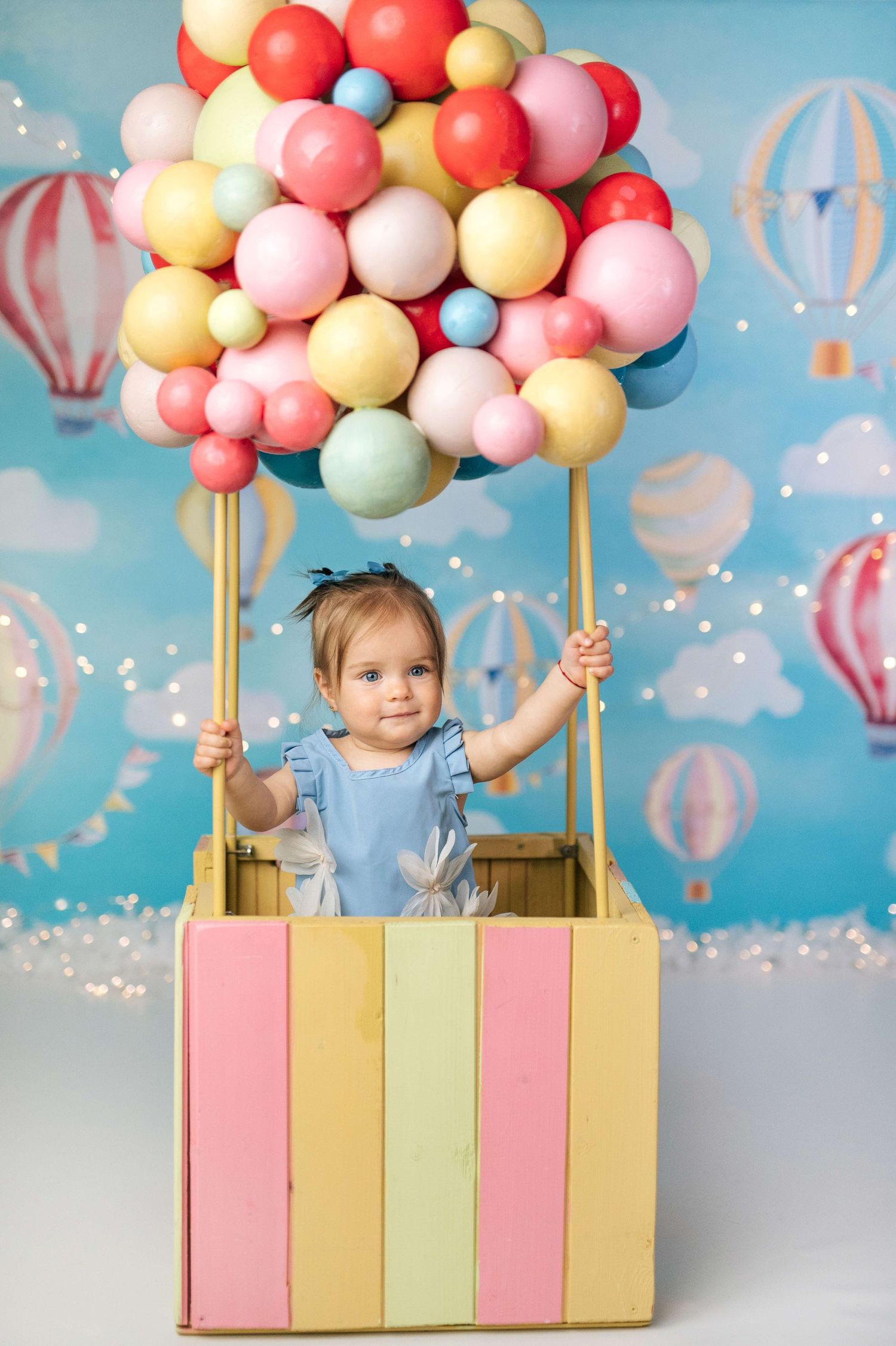Toddler girl in blue dress sitting in colorful striped hot air balloon prop with pastel balloons on sky backdrop.