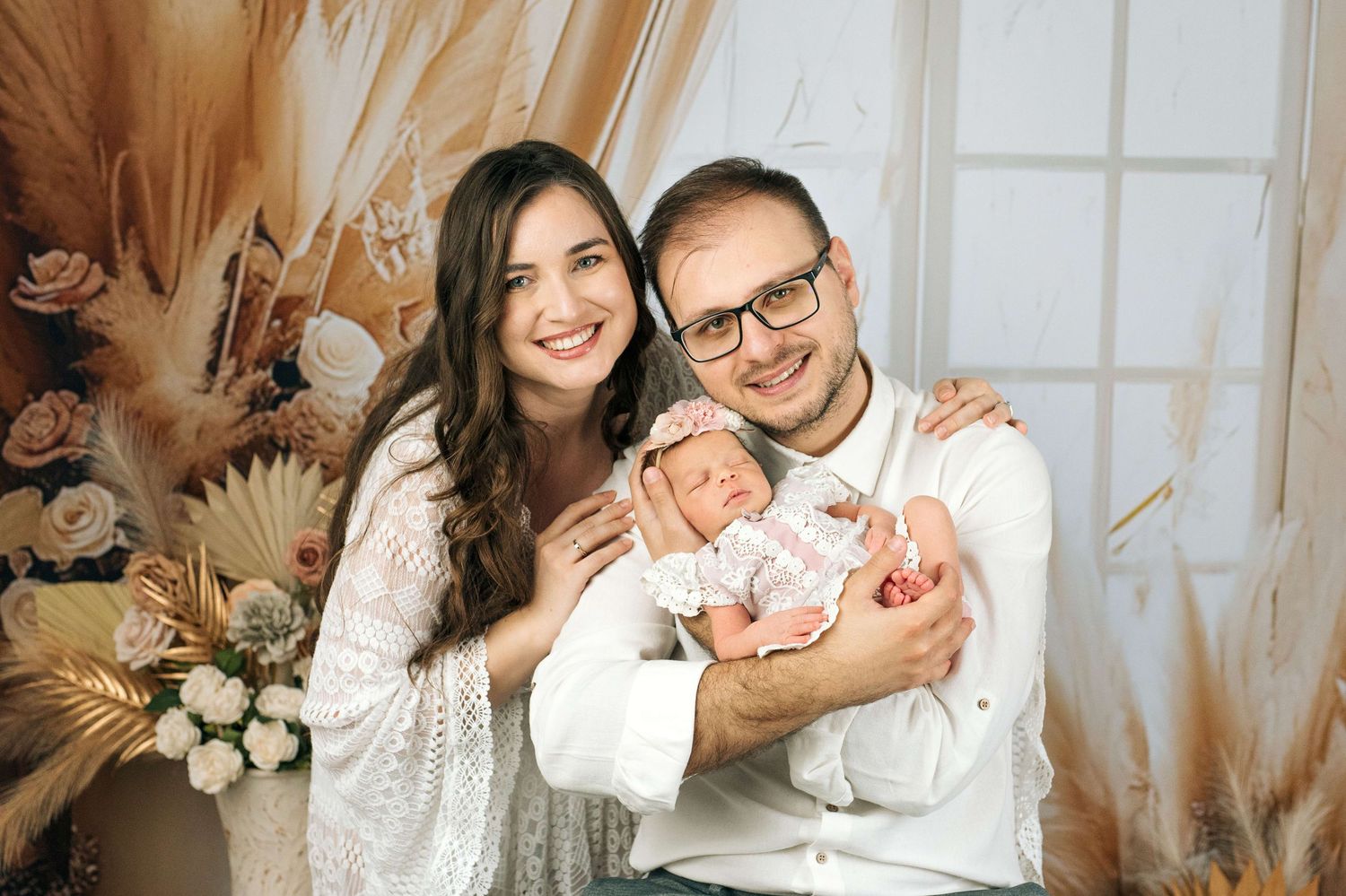 Happy couple smiling and holding their newborn baby in a warm, floral-decorated studio portrait setting.