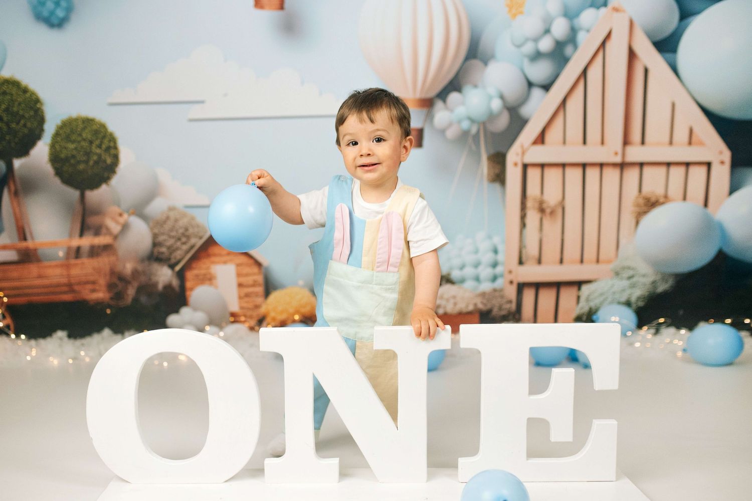 Toddler boy in blue overalls celebrating first birthday with large ONE letters and blue balloons in decorated studio.