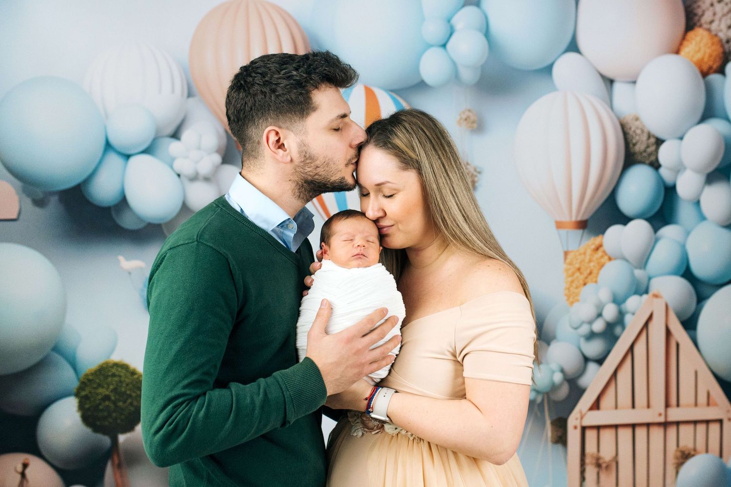 New parents sharing a tender moment kissing their newborn baby in a dreamy blue balloon backdrop studio photo.