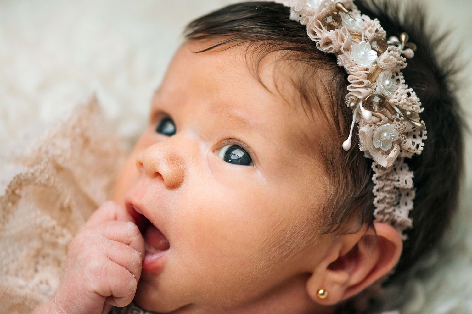Adorable newborn baby girl with dark hair wearing a floral headband and gold earring, fingers in mouth.