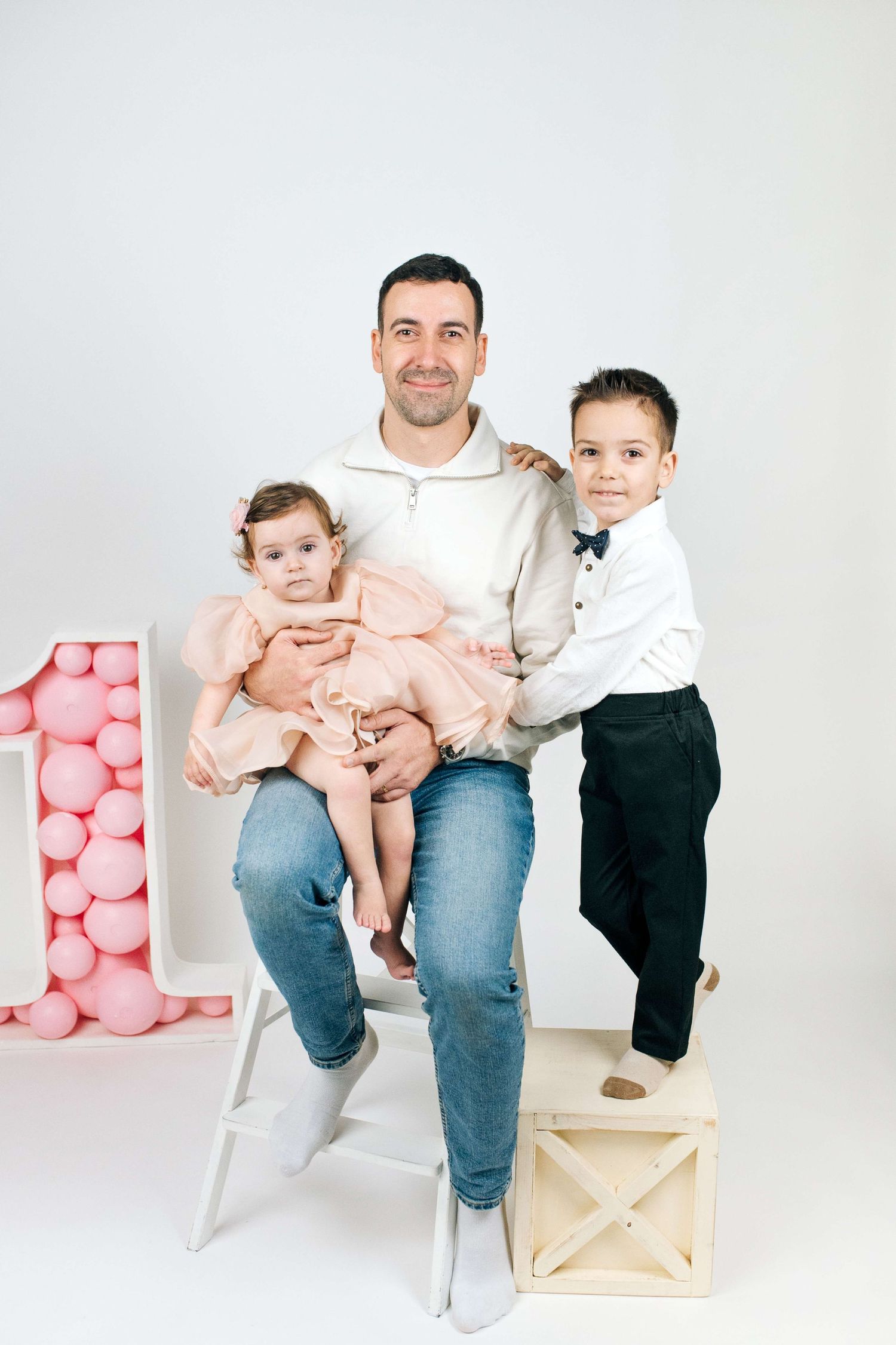 Father sits holding baby girl in pink dress while young boy stands beside them at a first birthday celebration.
