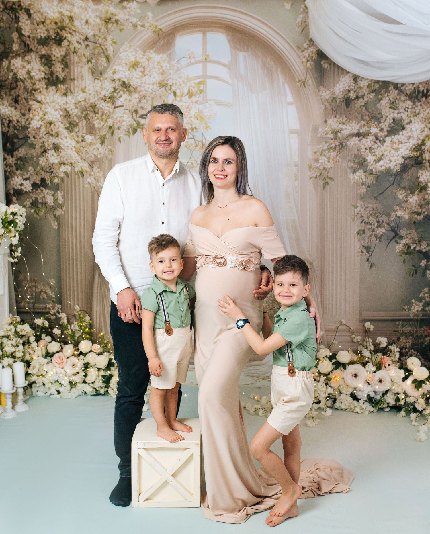 Pregnant woman in beige gown poses with husband and two young sons in green shirts amid white floral backdrop.