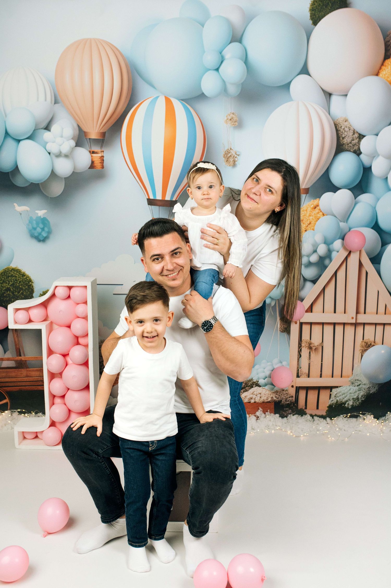 Family of four posing at a first birthday party with hot air balloon decorations and colorful balloons backdrop.