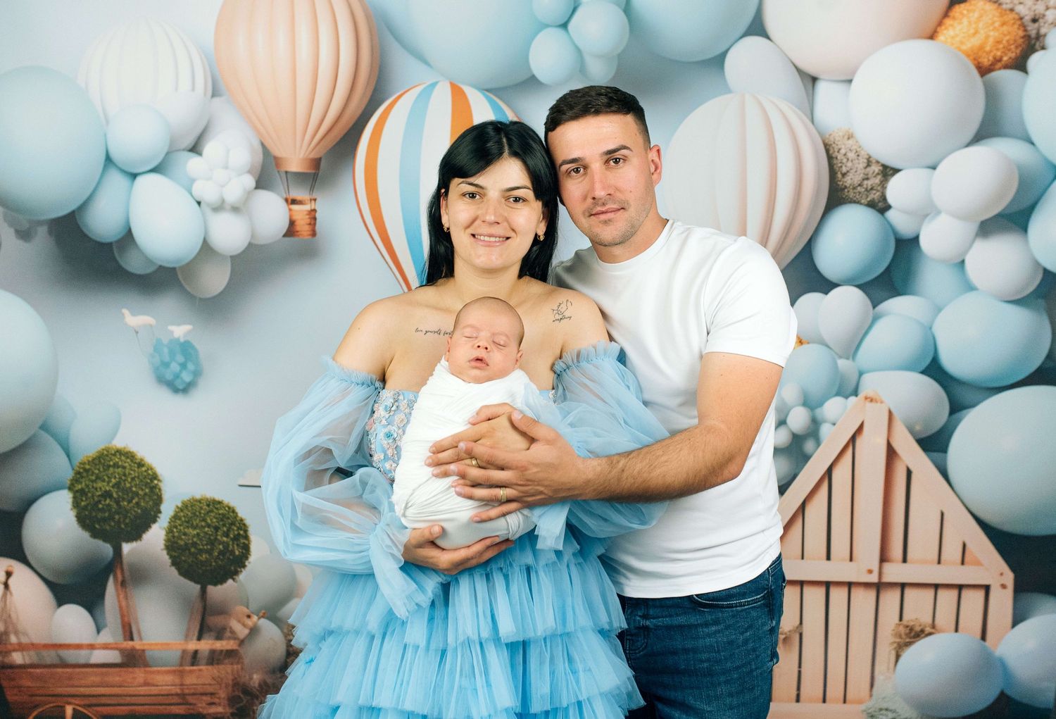 Young couple holding newborn baby in blue tulle dress, posing in front of blue balloon and hot air balloon backdrop.