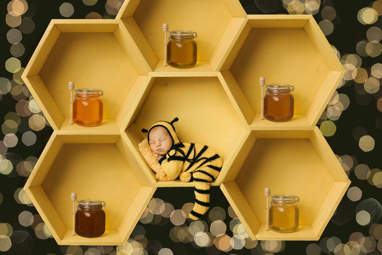 Newborn baby in bee costume sleeping in yellow honeycomb shelf with honey jars and bokeh lights.