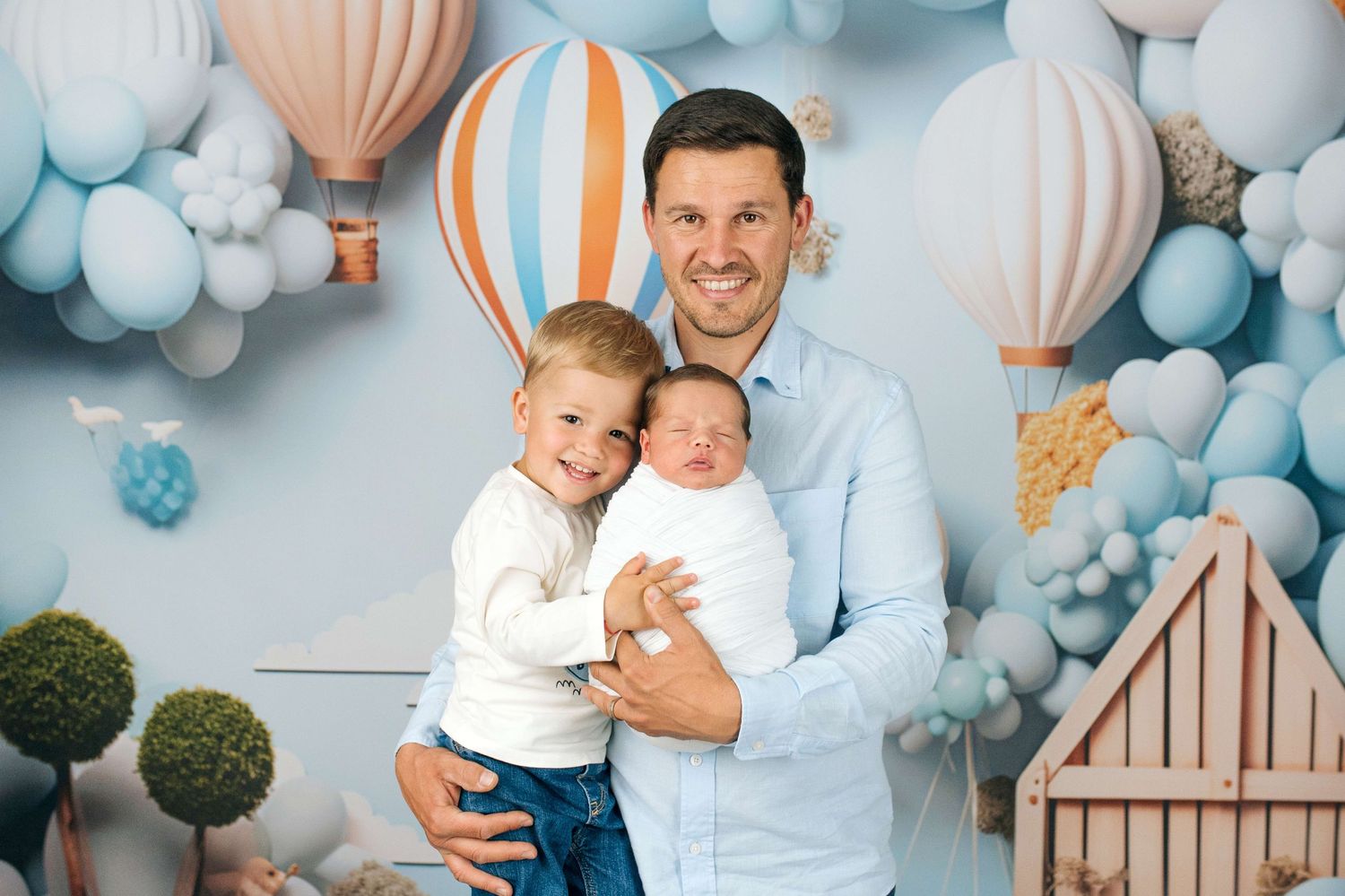 A father holds a newborn baby while posing with his toddler son in front of a blue balloon and hot air balloon backdrop.