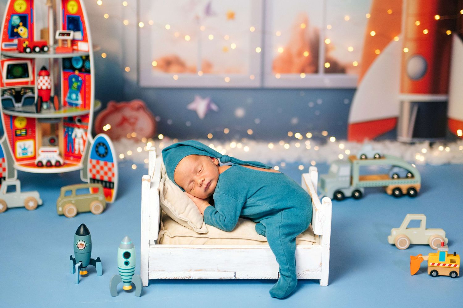 Newborn baby sleeping peacefully in blue onesie on white miniature bed surrounded by colorful toy rockets and cars.