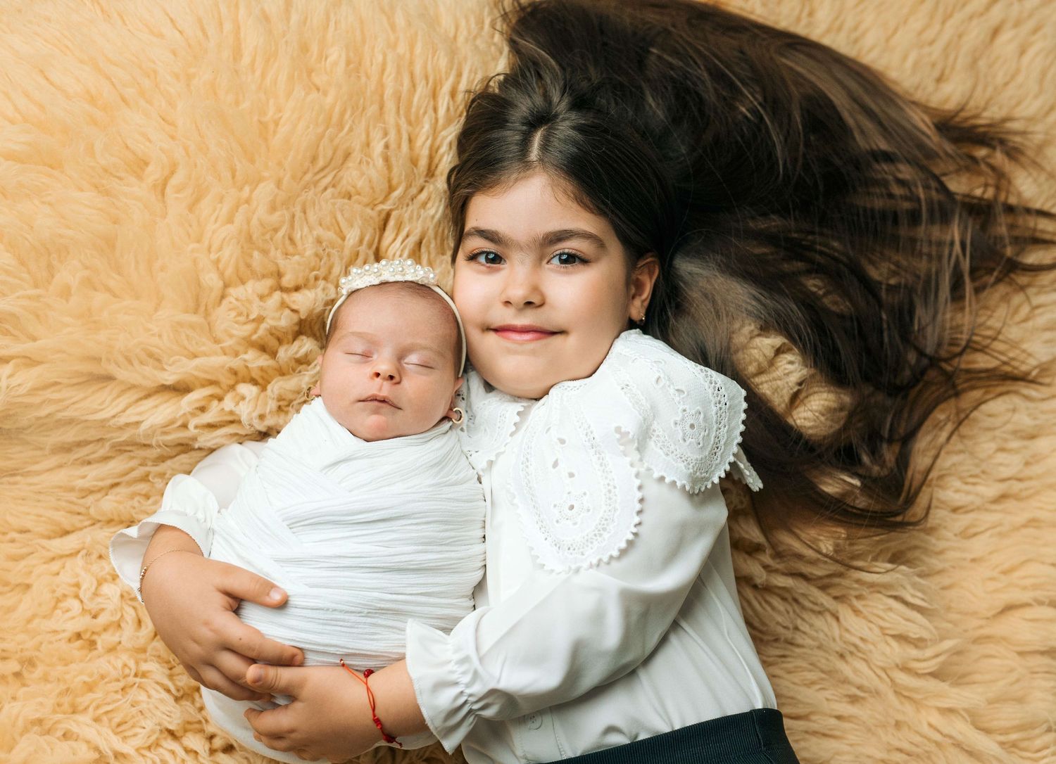 Older girl with long dark hair lying on fur rug holding sleeping newborn baby wrapped in white.