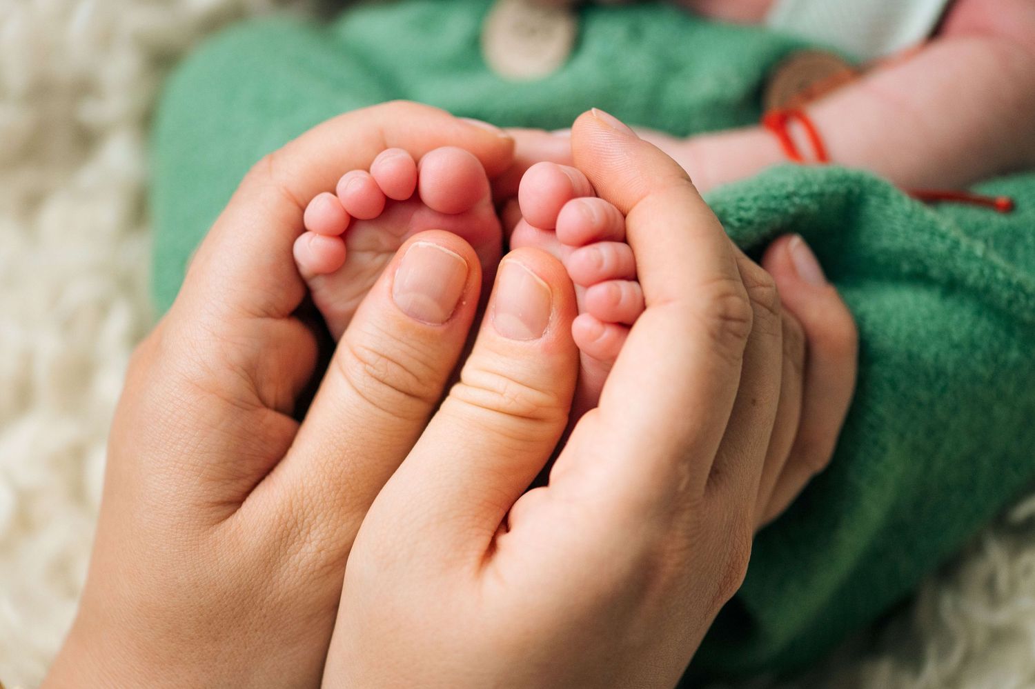 Adult hands gently cradling tiny newborn baby feet, showcasing delicate infant toes against a soft green blanket.