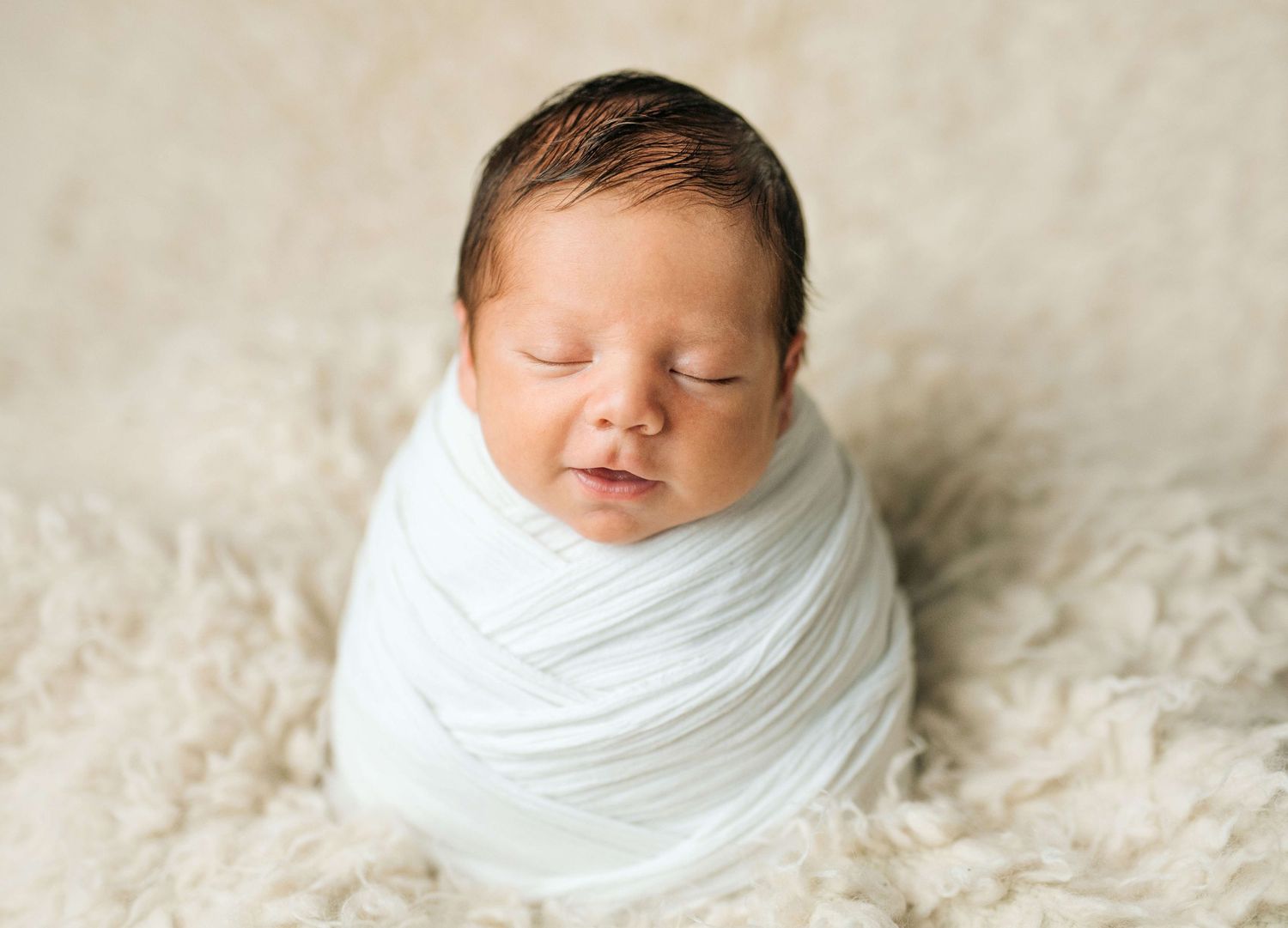 Newborn baby swaddled in white wrap sleeping peacefully on a fluffy cream rug in a studio photo.