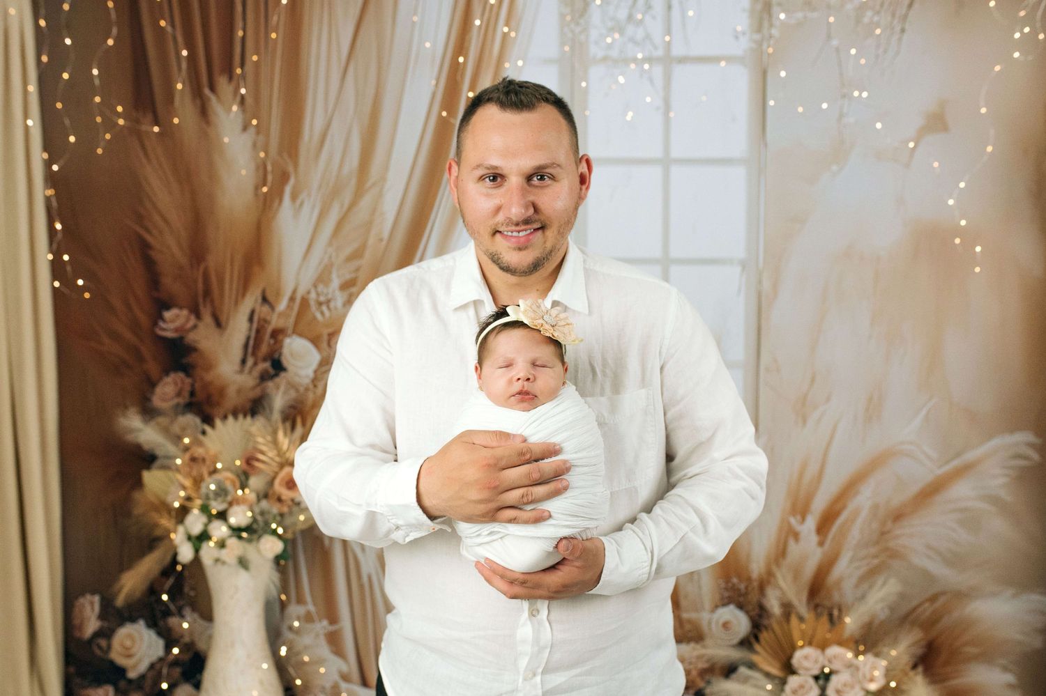 A man in a white shirt holds a swaddled newborn baby in a boho-styled photo studio with floral decor.