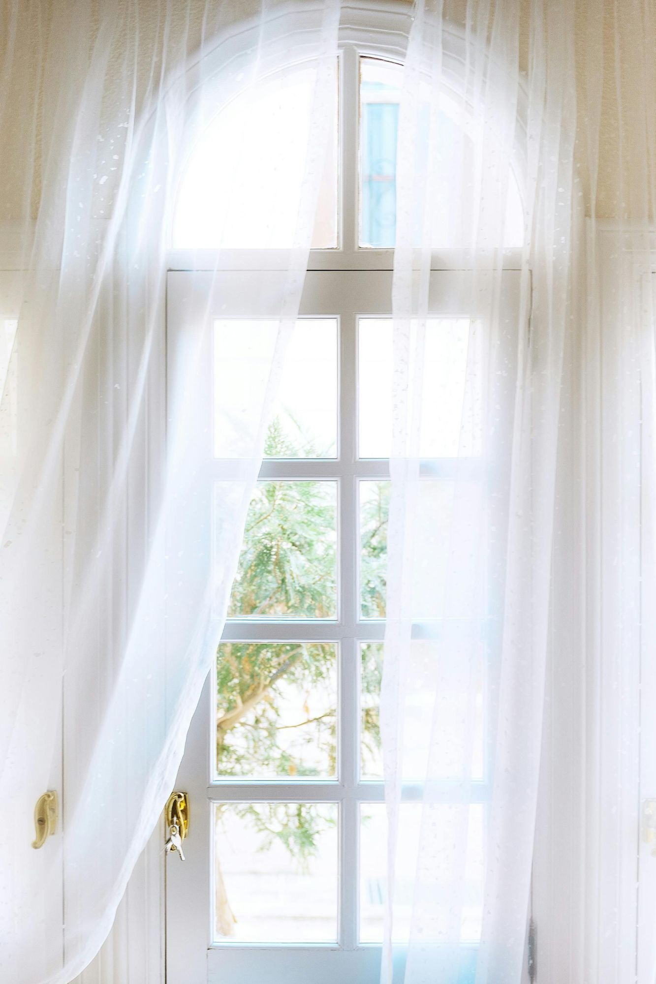 Sheer white curtains billowing in the breeze by an open window with natural light and green trees outside.