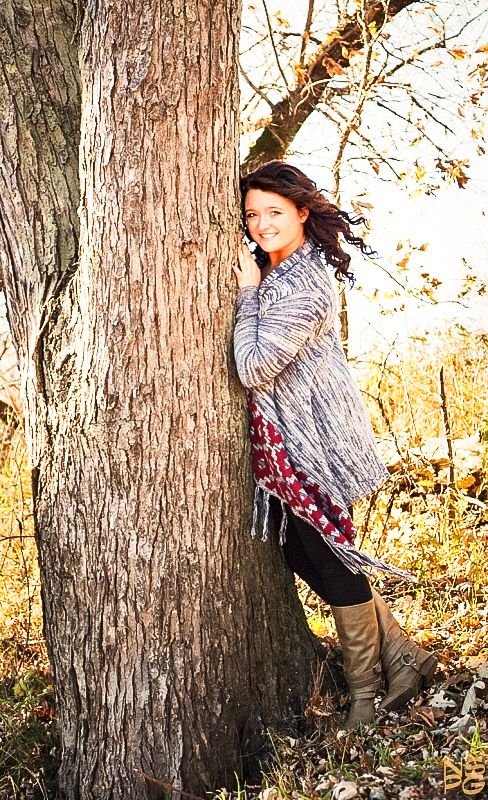 Young woman in gray wrap and boots leans against a large tree trunk in autumn foliage, smiling warmly.