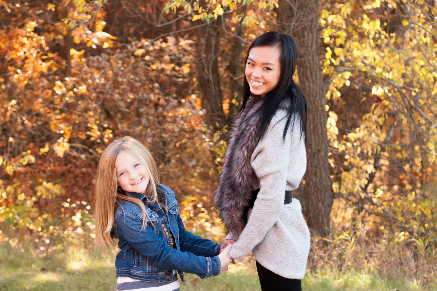 Two girls holding hands and smiling in an autumn forest, wearing cozy fall outfits among colorful orange and yellow foliage.