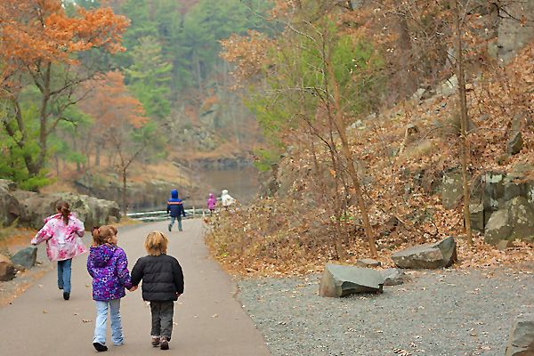 Children walk hand-in-hand along a scenic autumn park path surrounded by colorful fall foliage and rocky terrain.