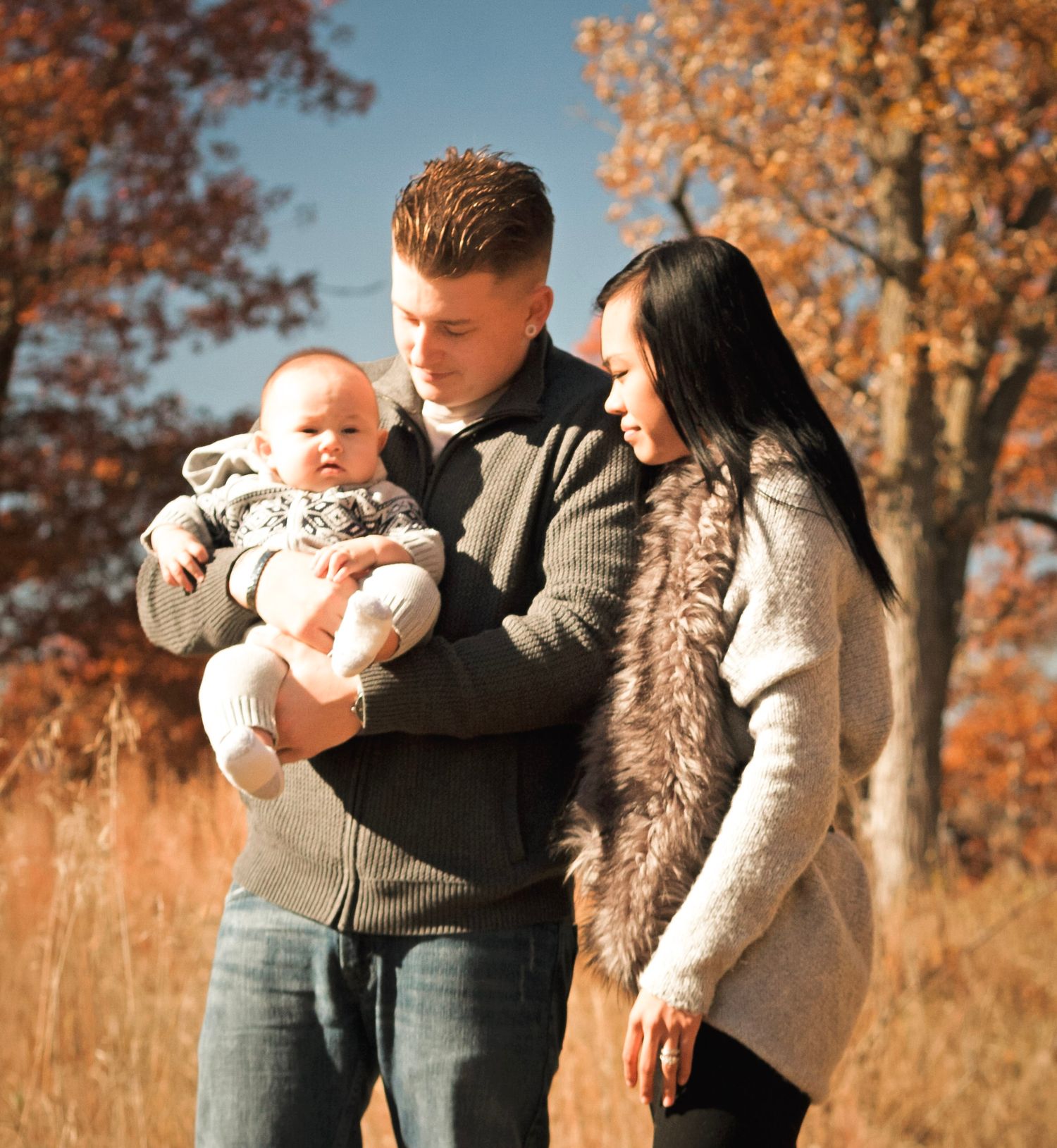 Young family with baby enjoying autumn outdoors, parents holding infant among fall foliage trees on sunny day.