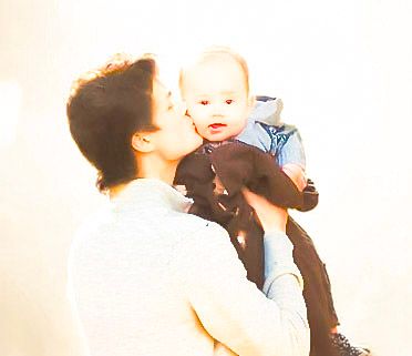 A person kisses a baby on the cheek while holding the infant up in a bright, softly lit room.