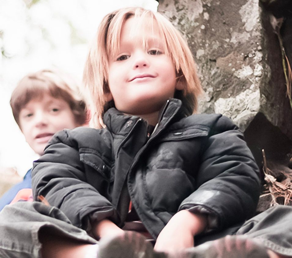 Two young children sitting outdoors near a stone wall, wearing jackets, smiling in a natural setting.