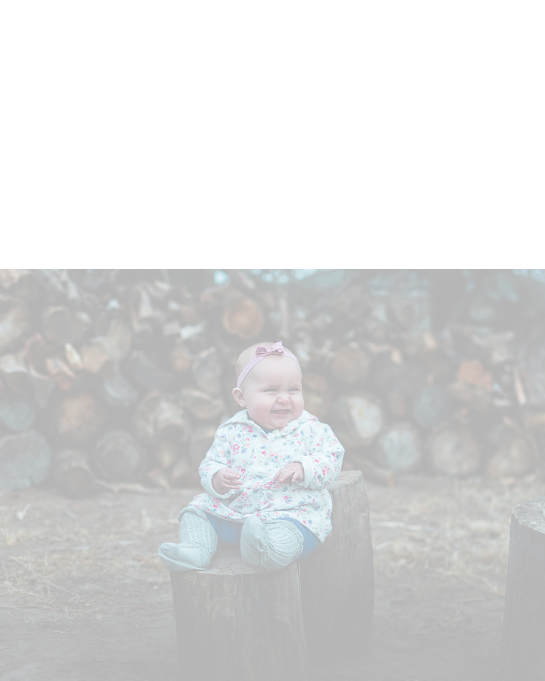 A happy baby girl in a floral outfit sits on a tree stump outdoors with stacked logs in the background.