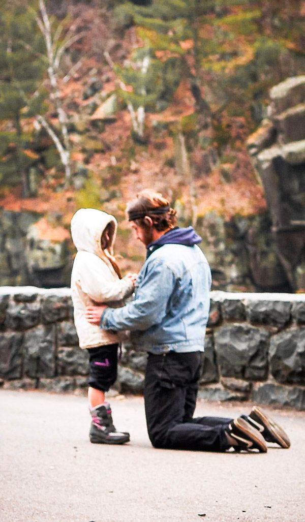 Adult kneeling on pavement to speak with young child in hooded jacket outdoors near stone wall in autumn.