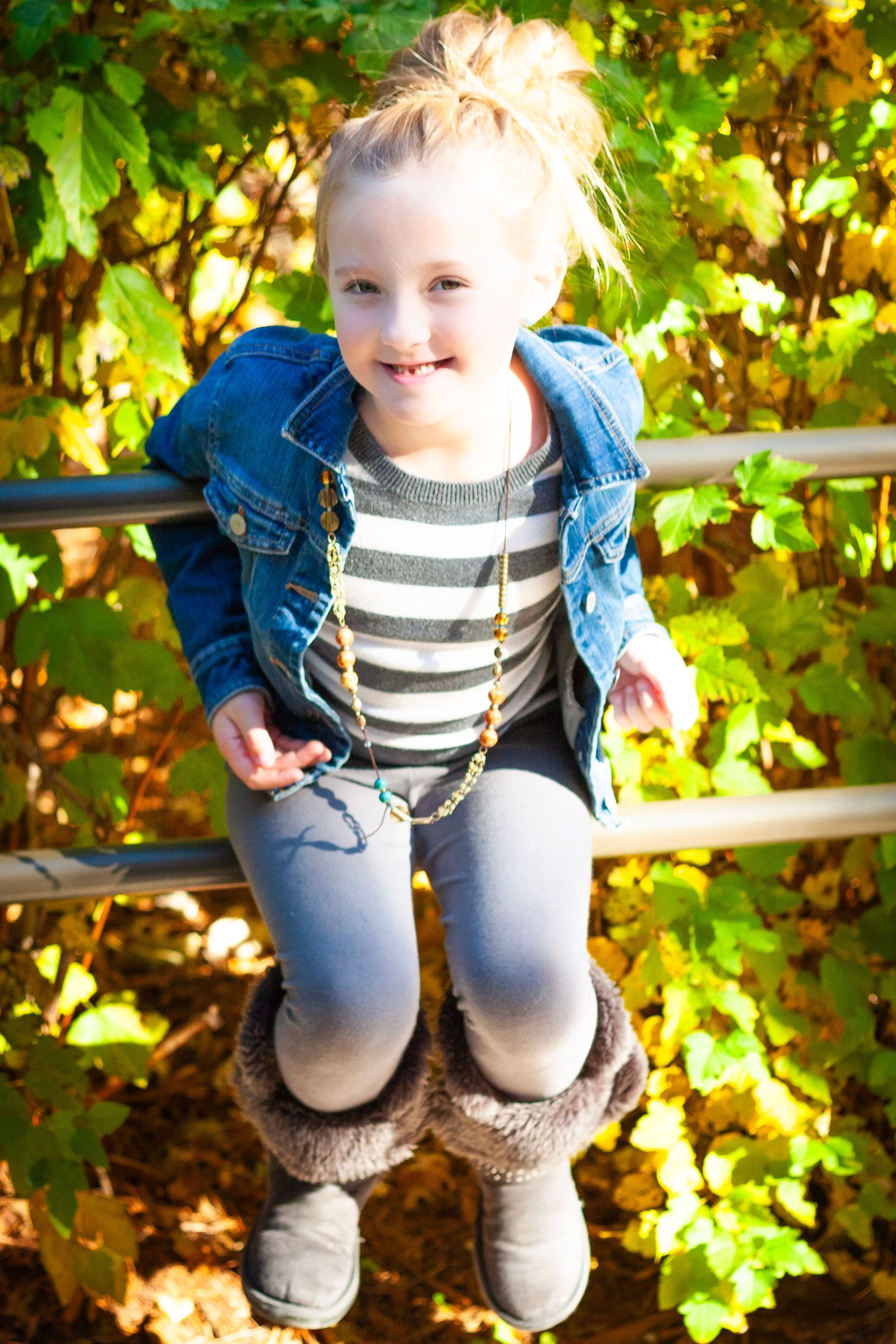 Young girl in denim jacket and striped shirt sitting on a fence rail surrounded by autumn foliage.