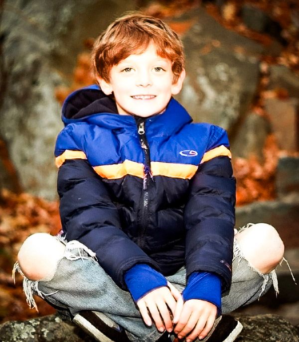 Smiling red-haired boy in blue jacket sitting cross-legged on rocks surrounded by autumn foliage.