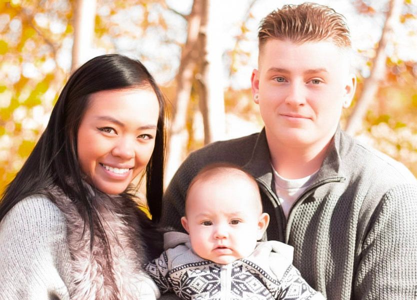 A smiling couple poses outdoors in autumn with their baby, wearing cozy sweaters amid golden fall foliage.