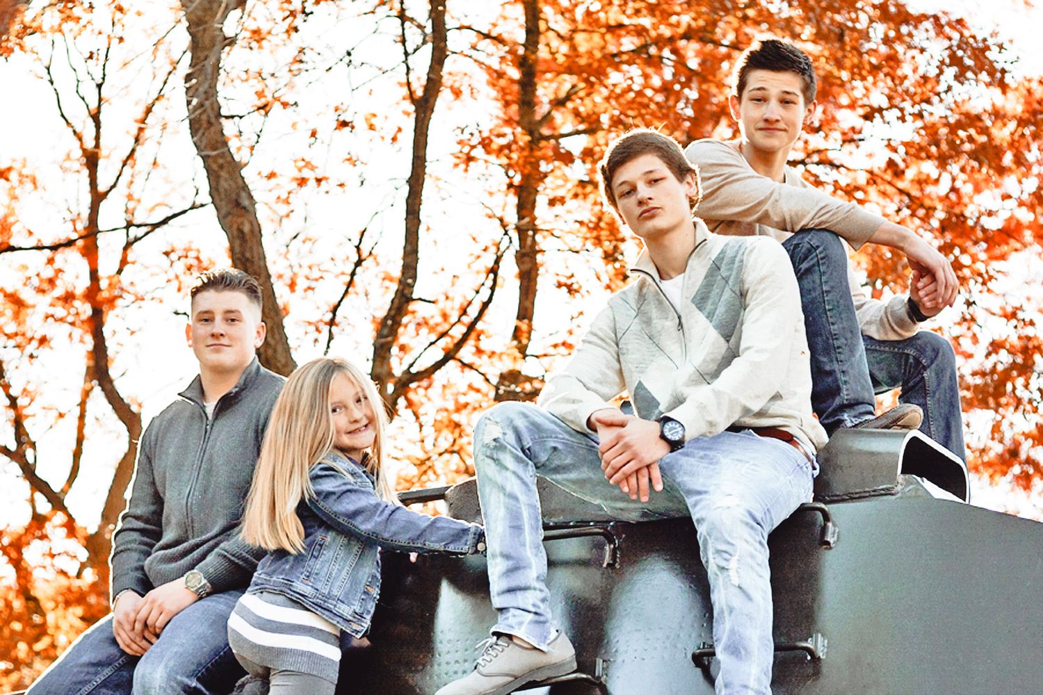 Four teenagers sitting on a military tank outdoors with vibrant autumn foliage in the background.