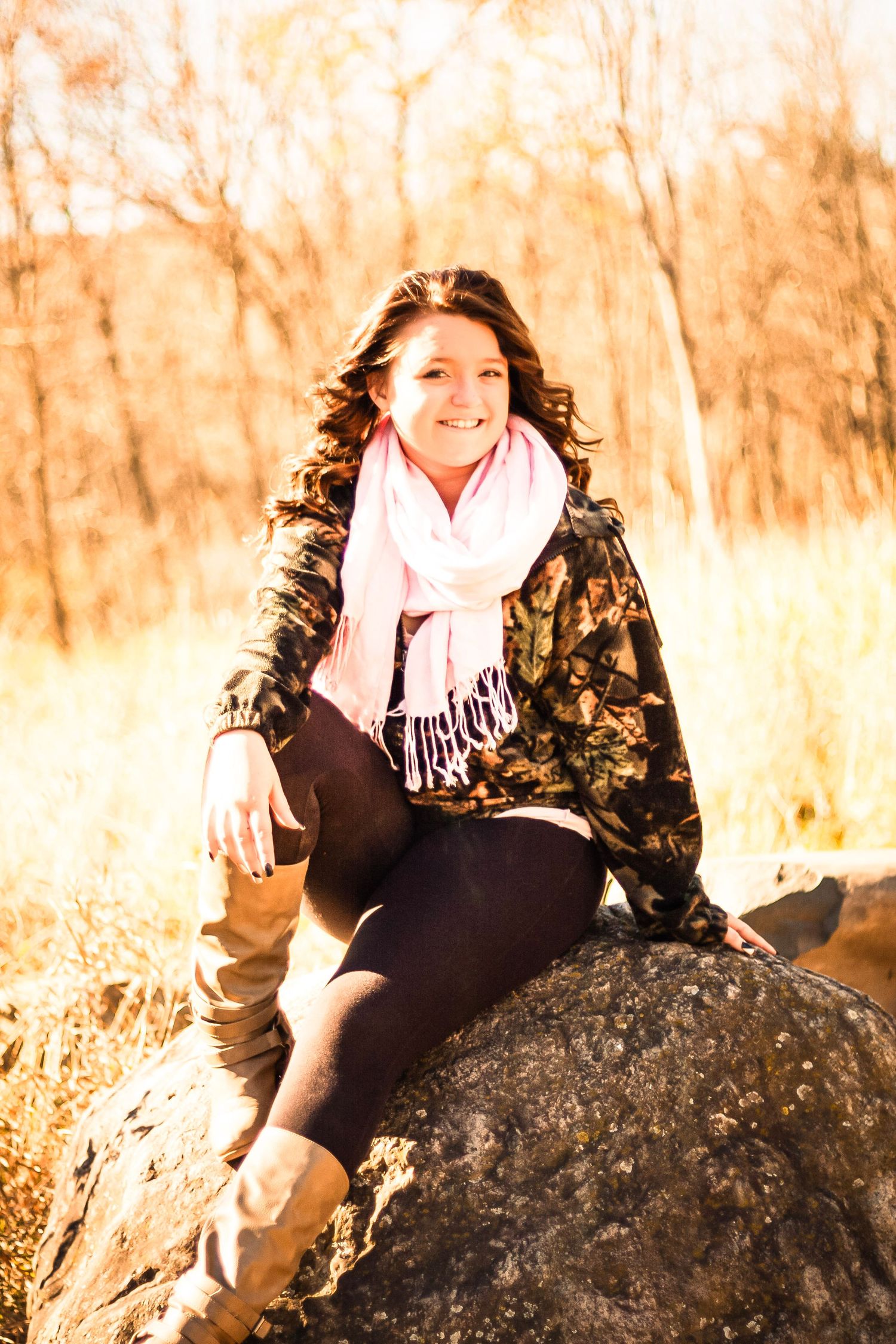 Smiling young woman in camo jacket and pink scarf sitting on a rock in an autumn forest setting.