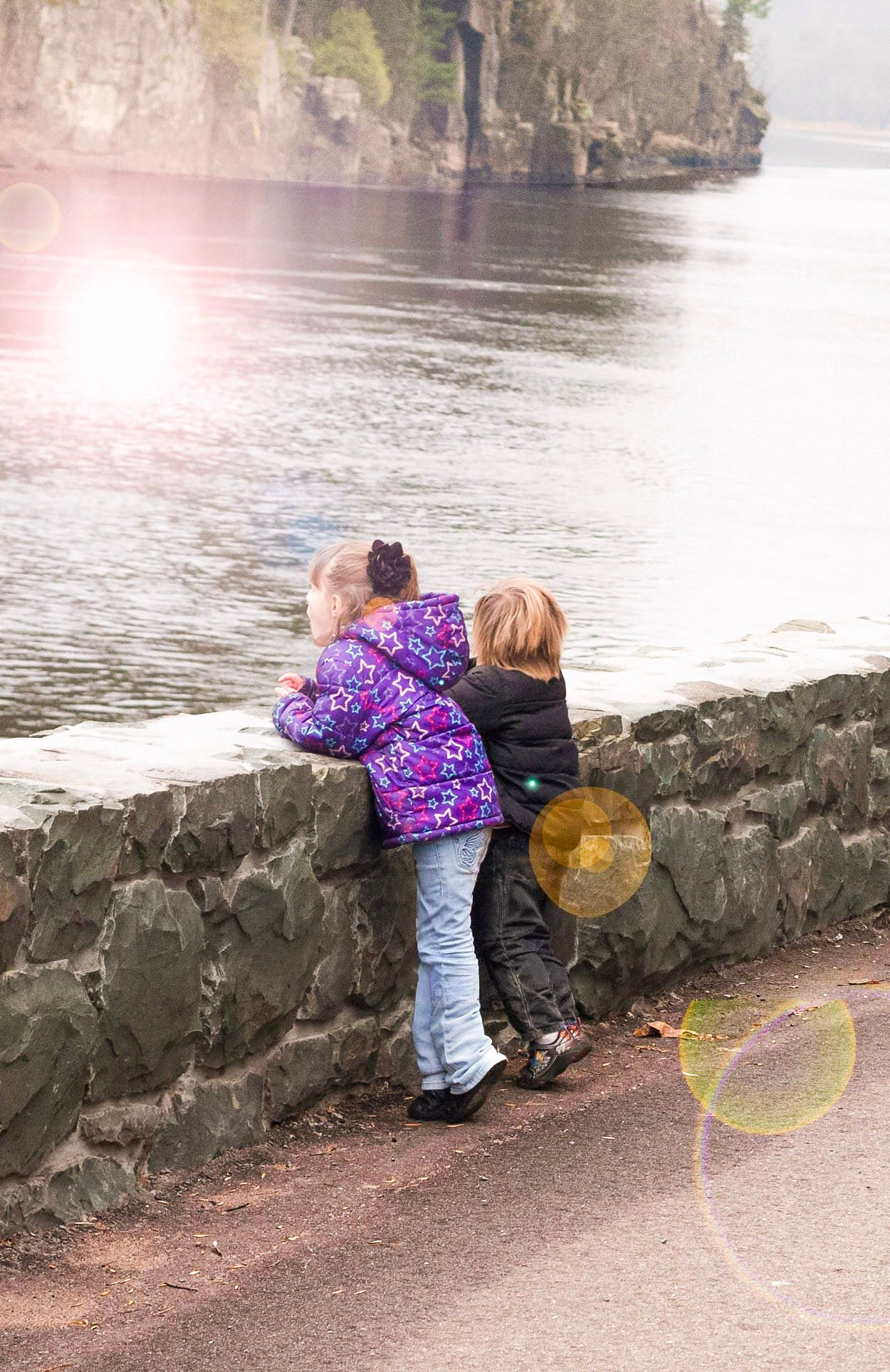 Two children in jackets lean over a stone wall to look at a scenic river with rocky cliffs in the background.
