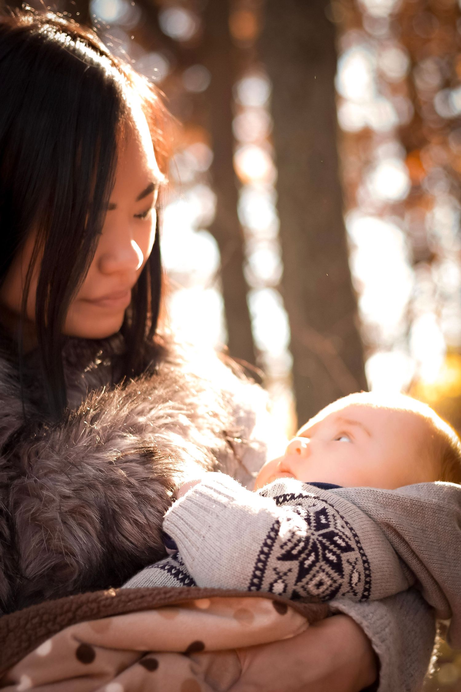 A young Asian mother tenderly holds her newborn baby outdoors in autumn sunlight, sharing a gentle moment.